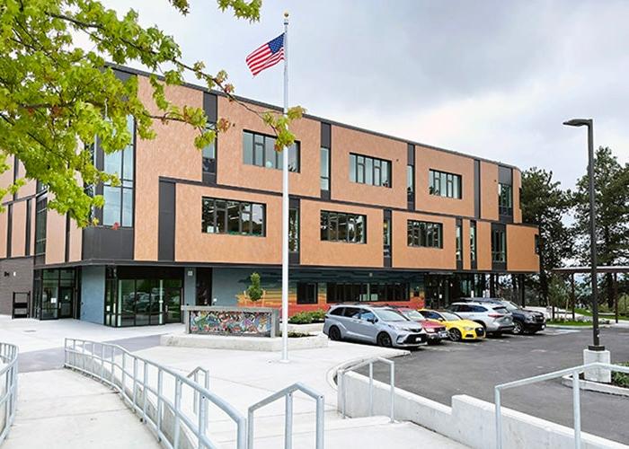 a 3 story building made of reddish bricks with the 2nd floor forming a canopy over the first floor entrance. a concrete walkway and parking lot are in front. there is an American flag on a pole.